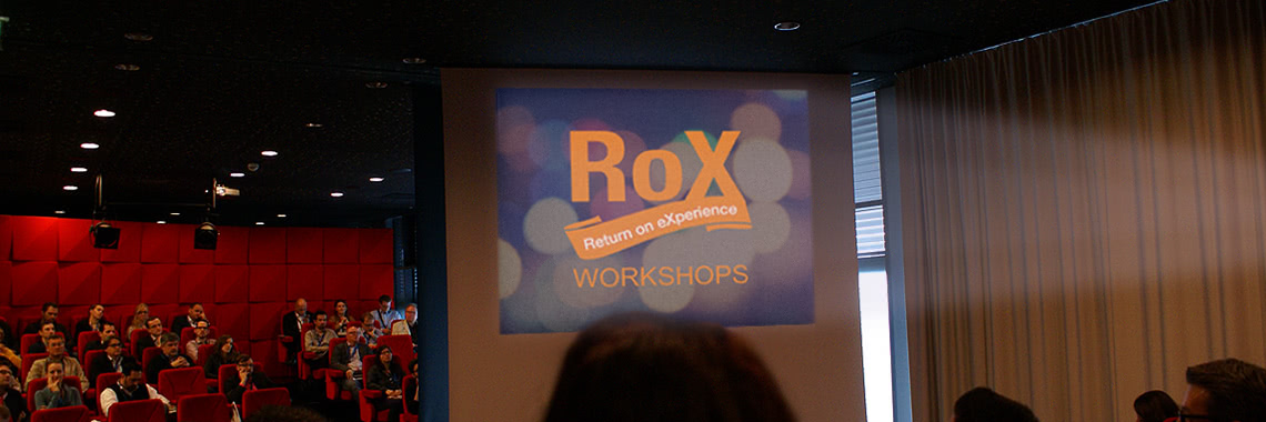 Saal mit Zusehern, Leinwand in der Mitte mit RoX Workshops Logo
