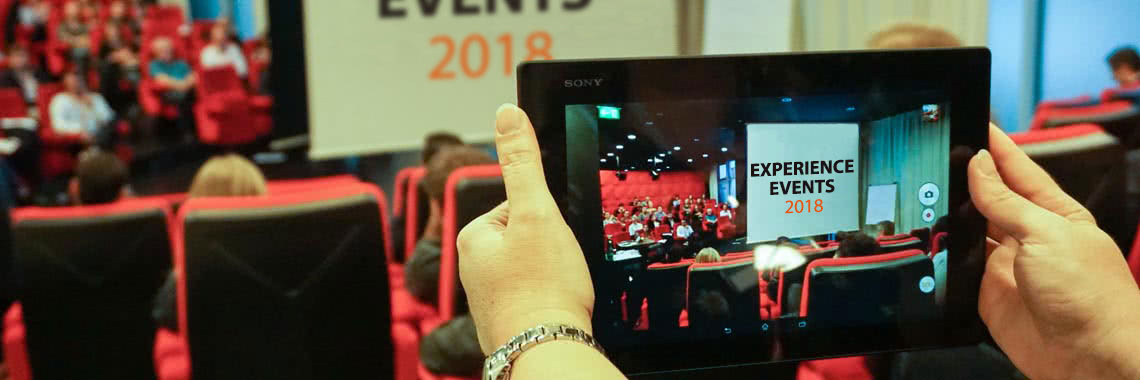 Roter Saal mit Publikum, im Vordergrund Haende die ein Tablet halten. Am Screen steht Experience Events 2018